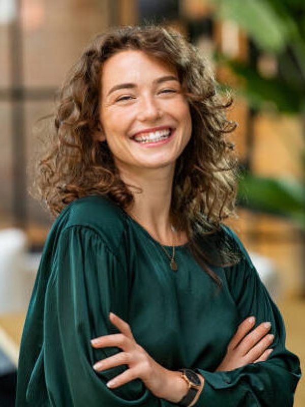 Portrait of young smiling woman looking at camera with crossed arms. Happy girl standing in creative office. Successful businesswoman standing in office with copy space.